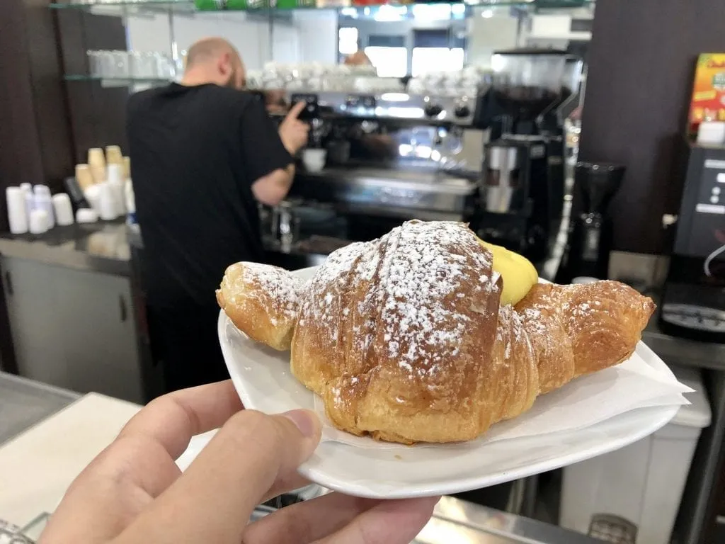 A cornetto crema being held up in a bar in Italy, with a man making an espresso visible in the distance--when ordering coffee in Italy, consider adding a pastry to your order as well!