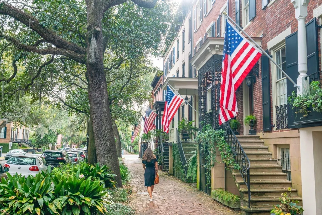 Kate Storm in a black dress walking away from the camera along Jones Steet in Savannah GA, with several American flags hanging from homes on the right side of the photo