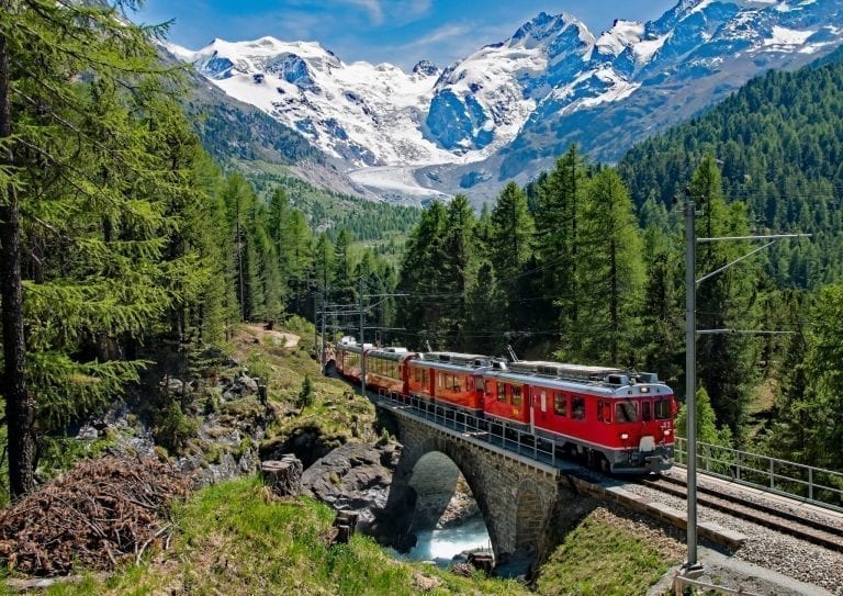 Red train moving through Switzerland with mountains visible in the background--views like this are one of the best reasons to travel Europe by train