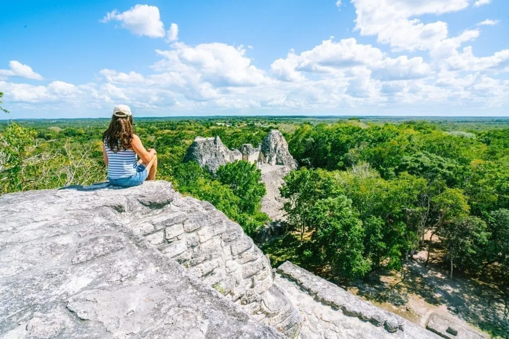 Kate Storm in a tank top and baseball cap sitting on a pyramid in Becan Mexico overlooking another structure