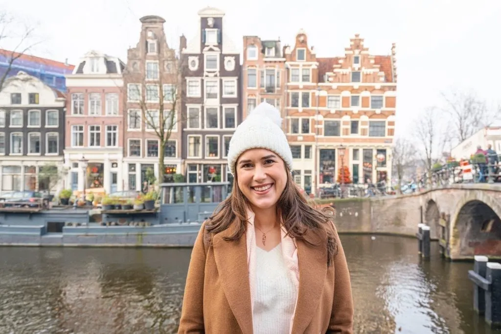 Kate Storm in a brown coat and white hat standing in front of a canal on a cloudy day in Amsterdam