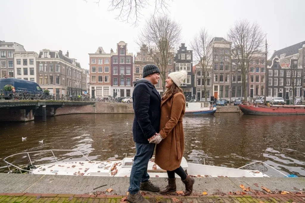 Kate Storm and Jeremy Storm wearing winter coats on the edge of a canal in Amsterdam in December, facing each other