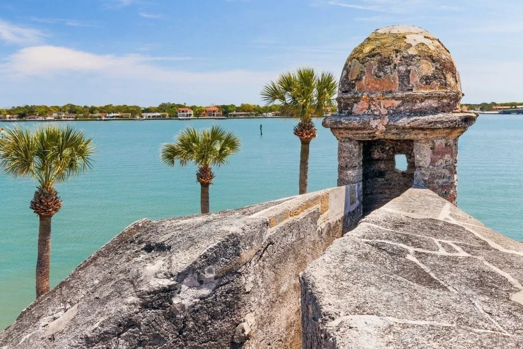 View of palm trees and ocean water over the stone wall of St Augustine Florida's 17th century fort. St Augustine is one of the best weekend trips in the south