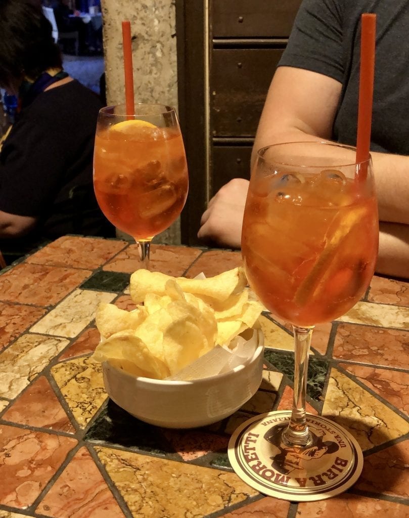 2 aperol spritzes on a small table in Rome at night with a small bowl of potato chips between them