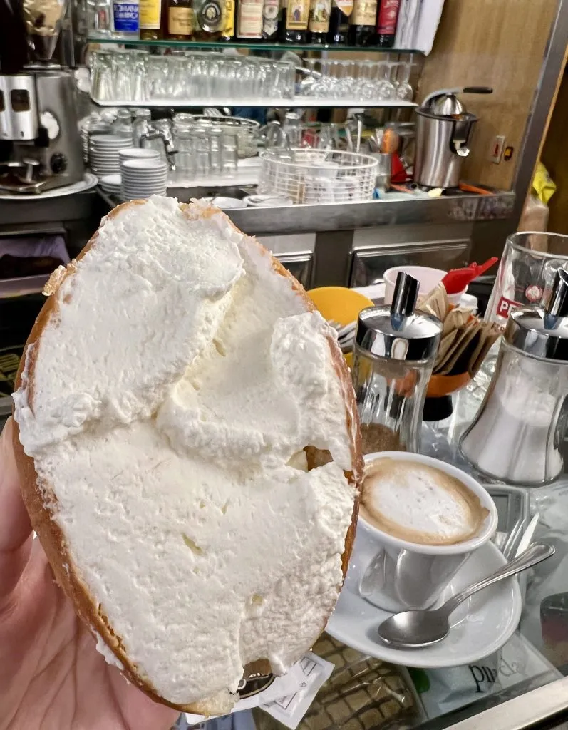 maritozzi being held up in an italian coffee bar in rome with cappuccino visible in the background
