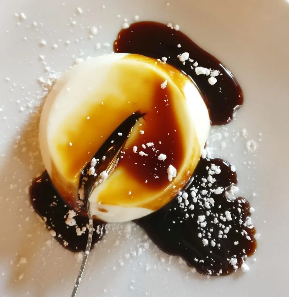 Round panna cotta with a spoon inserted in it shot from above. The panna cotta is coated in caramel sauce. Panna cotta is a delicious dessert to consider when deciding what to eat in Rome Italy