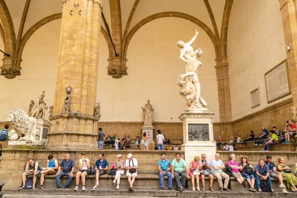 Tourists sitting in front of statues displayed in Piazza Signoria, as seen as part of a Florence itinerary