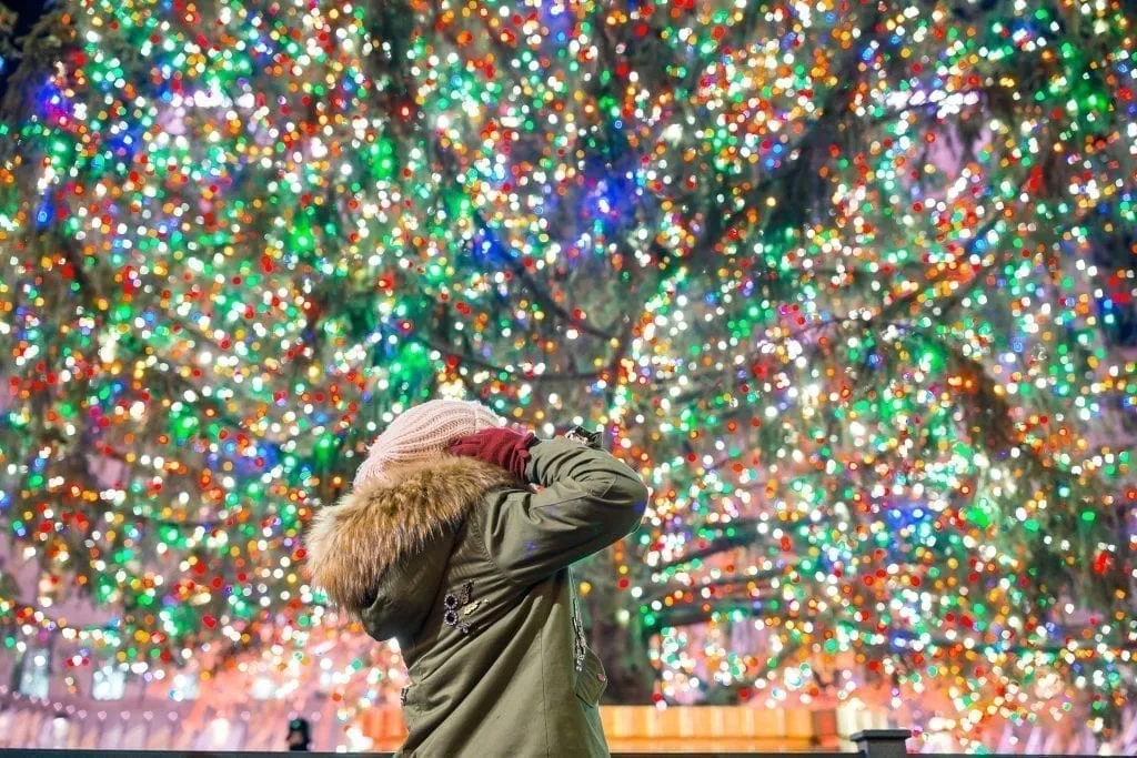 Woman in a green coat looking up at the Rockefeller Christmas tree with colorful lights. NYC is a classic Christmas vacation usa