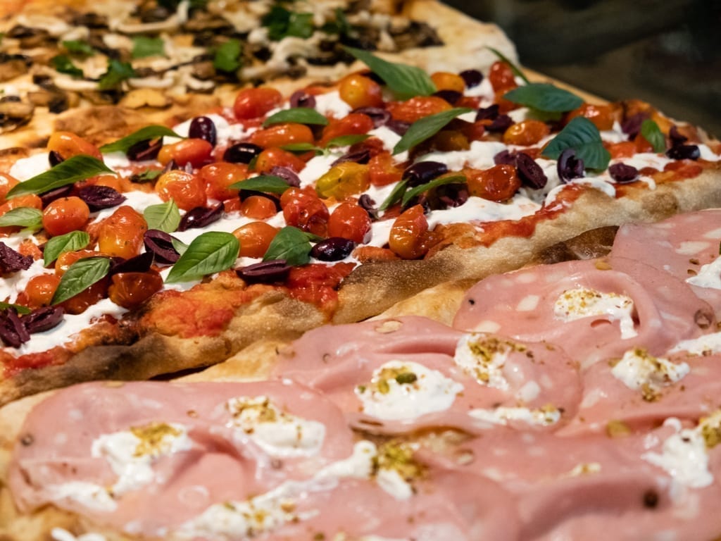 Pizza al Taglio laid out in pans, with a mortadella flavor in the foreground and a tomato and olive pizza in the background. When looking for the best food in Rome, be sure to try pizza al taglio!