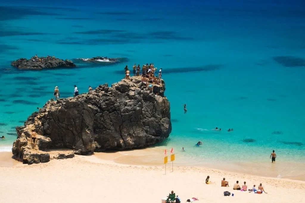 Waimea Beach in Oahu Hawaii with a large rock on the left side of the shot and people sitting on both the rock and sand