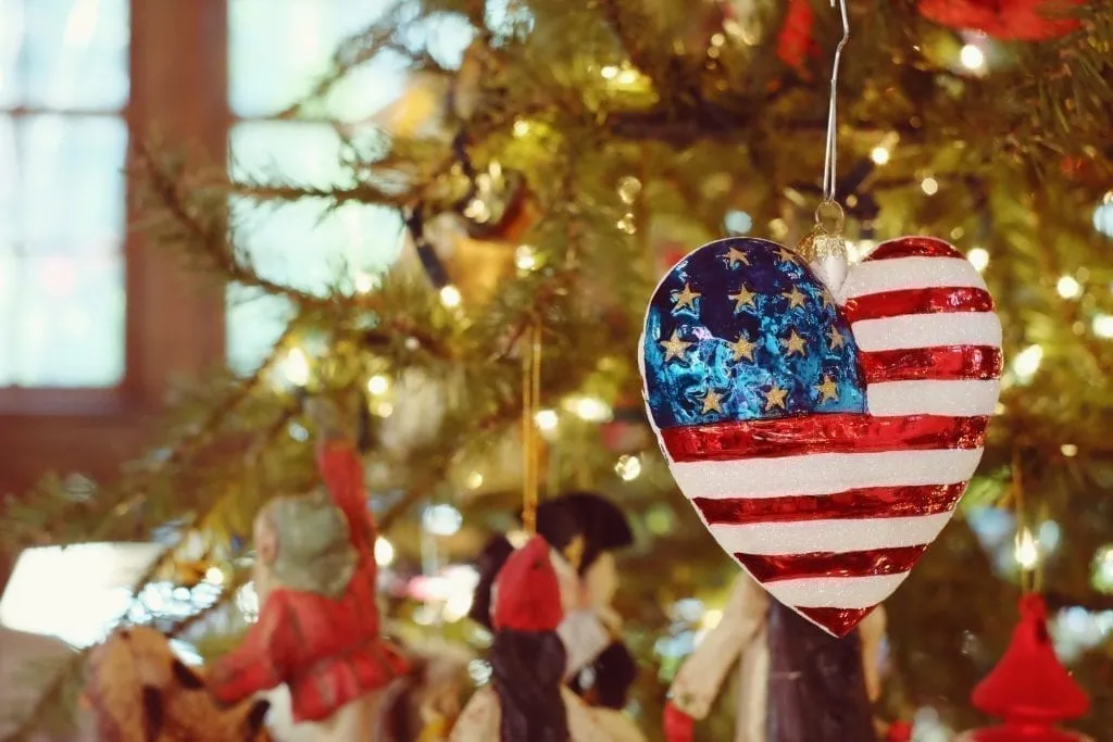 Heart-shaped Christmas ornament with an American flag painted on it hanging from a tree. This post rounds up the best christmas vacations in us