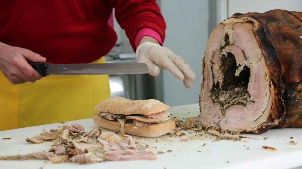 Porchetta sandwich in Rome with a large piece of porchetta off to the right and a man holding a knife over the sandwich