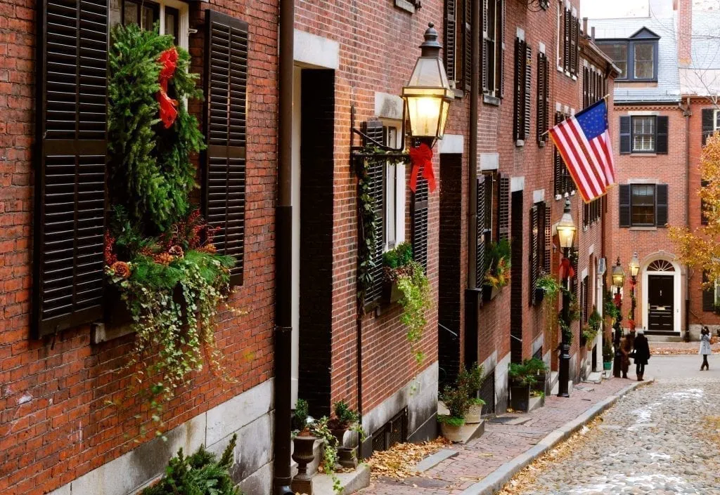 Acorn Street in Boston MA with Christmas decor hanging from the buildings on the left and an American flag in the distances. Boston is one of the best Christmas vacations in us