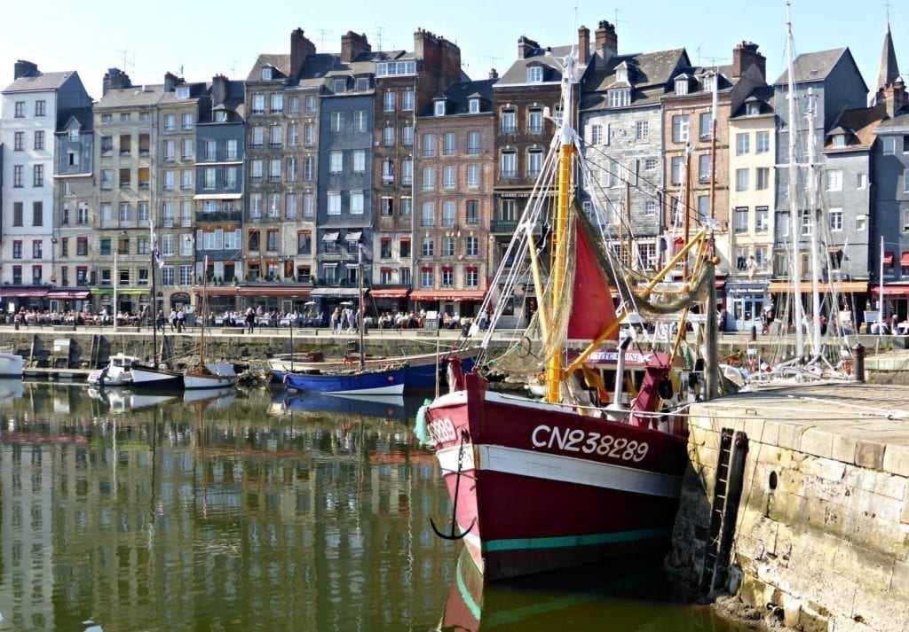 Harbor of Honfleur, one of the prettiest small towns in France. a small red boat is in the foreground and a line of buildings in the background