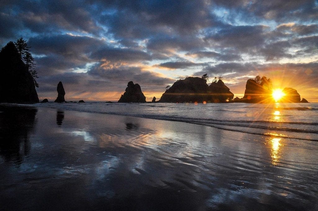 Shi Shi Beach at sunset, with dramatic clouds in the sky and a sunflare in the right corner of the photo
