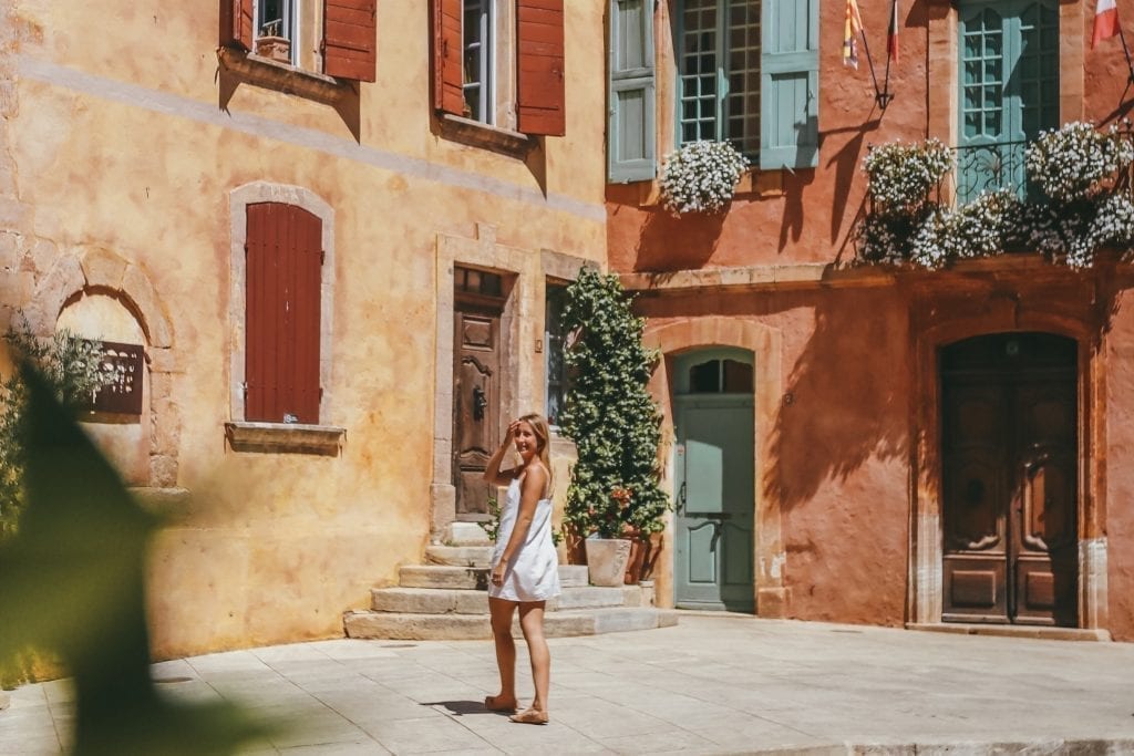 Photo of a blonde woman in a white dress standing in front of colorful red and yellow buildings in Roussillon