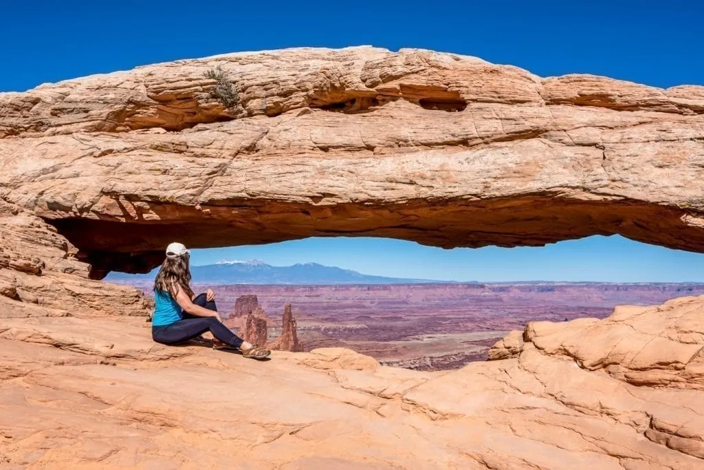 Kate Storm in a blue tank top sitting in front of Mesa Arch in Canyonlands National Park Utah