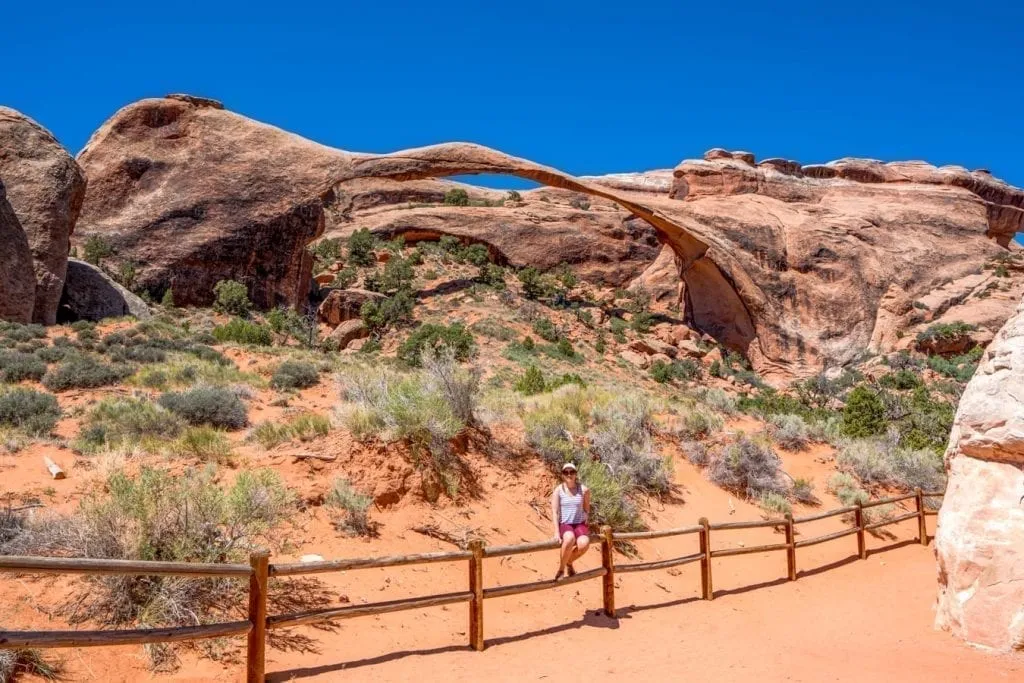Kate Storm sitting on a fence in front of Landscape Arch, one of the best hikes in Arches National Park