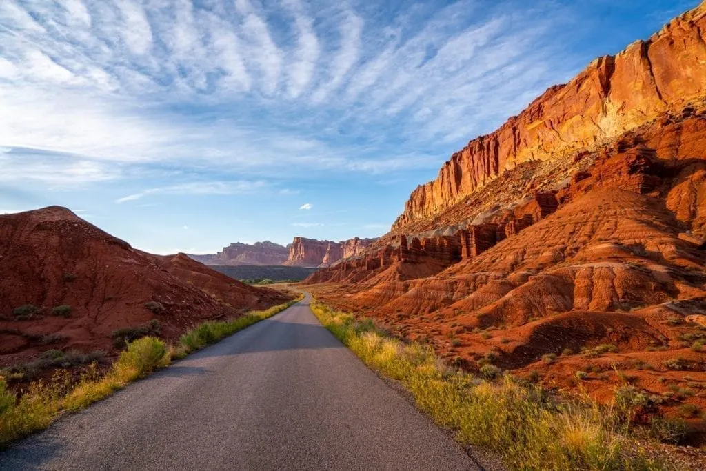 Empty road in Capitol Reef National Park near sunset with golden rock formations on either side of the road. Views like this are an essential part of road trips in Utah!