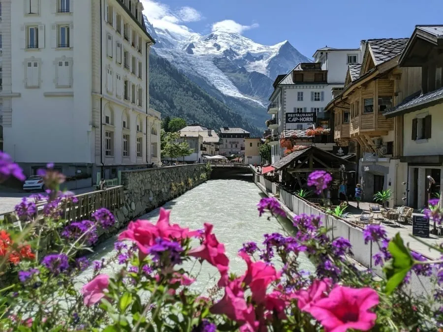 Photo of a canal in Chamonix in summer, with blooming pink flowers in the foreground and a snow-capped mountain in the background. Chamonix is one of the best small towns in France.