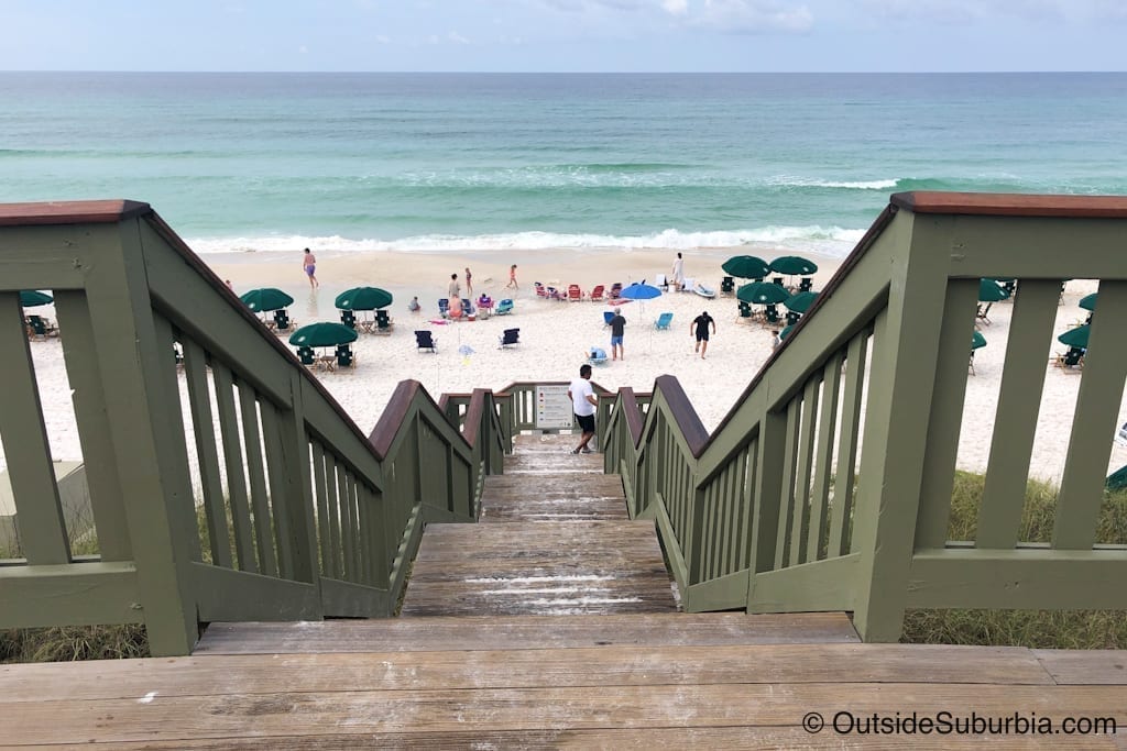 Wooden steps descending to a beach in South Walton Florida. Blue umbrellas are set up on the sand.