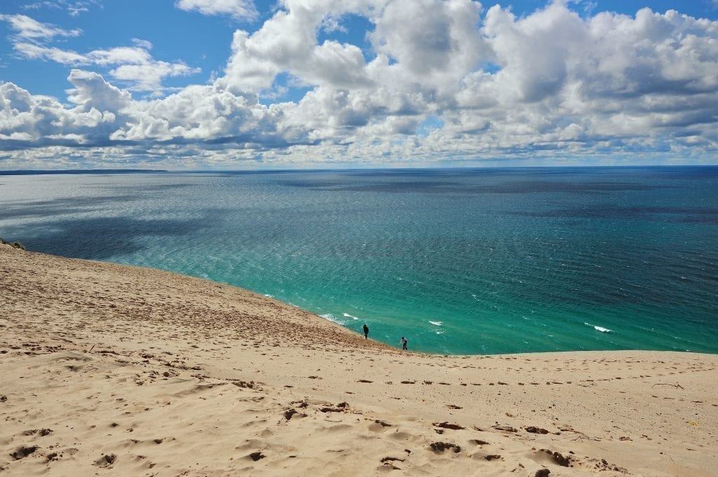 Sandy dunes of Lake Michigan with turquoise lake in the background and 2 small human figures in the distance. Lake Michigan is home to some of the best beaches in america