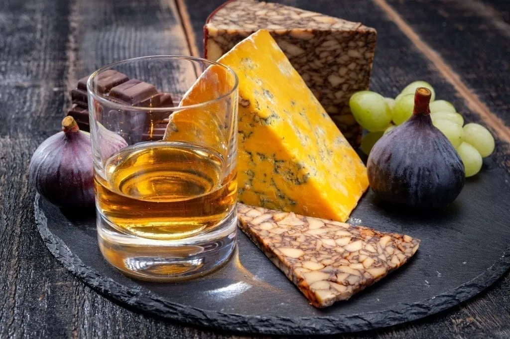 Plate of Irish cheese being displayed with a glass of whiskey on a black platter. Irish cheese is one of the best Ireland souvenirs