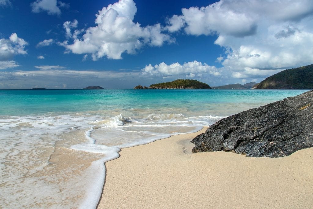 Bright blue water of Cinnamon Bay in St John on a sunny day, with golden sand in the foregeround and a black rock on the right side of the photo. This Caribbean beach is a beauty.