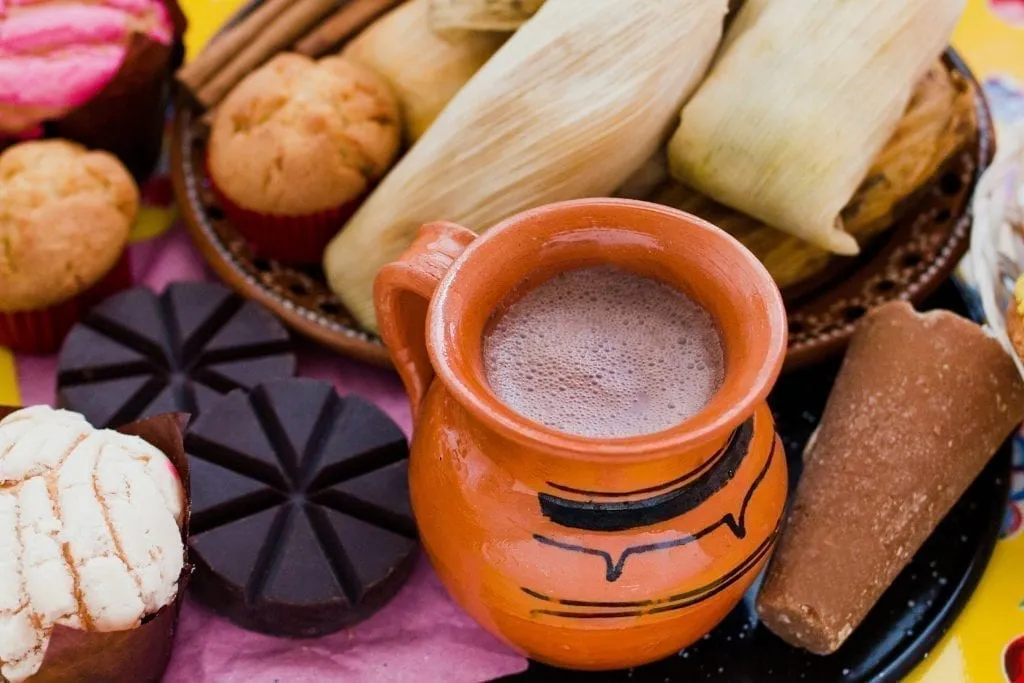 Cup of atole in an orange mug with tamales behind it and pan dulce to the left of it.