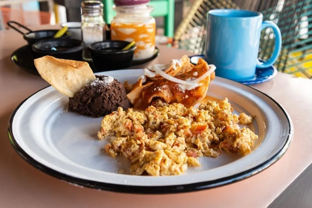 Huevos a la Mexicana on a white plate with a small serving of beans behind it and a blue mug in the back right of the photo