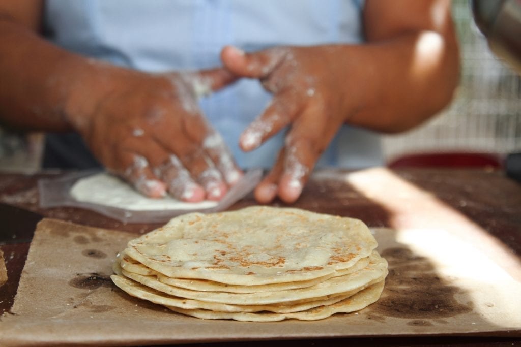 Stack of fresh corn tortillas in the foreground with the hands of a woman shaping another tortilla in the background