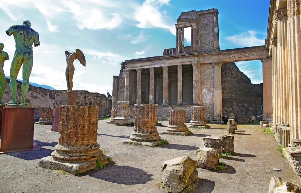 Photo of a square in Pompeii Italy with ruins of columns and several statues in it