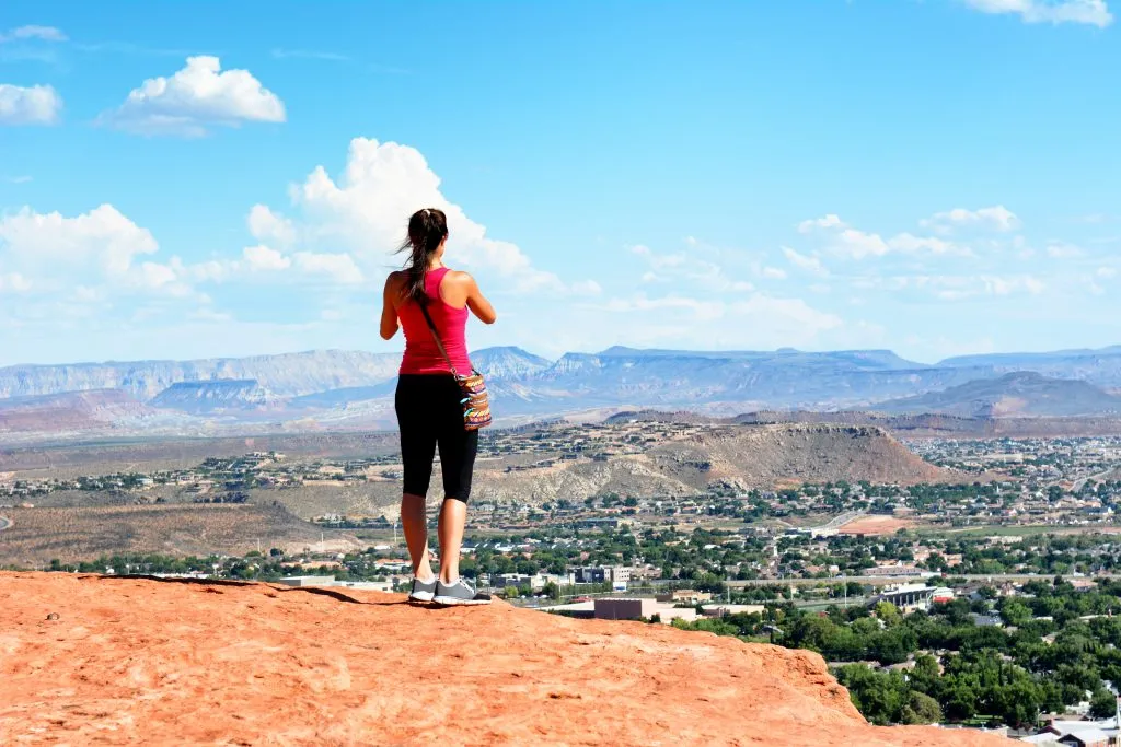 woman in a red tank top overlooking st george utah from above on a hiking trail