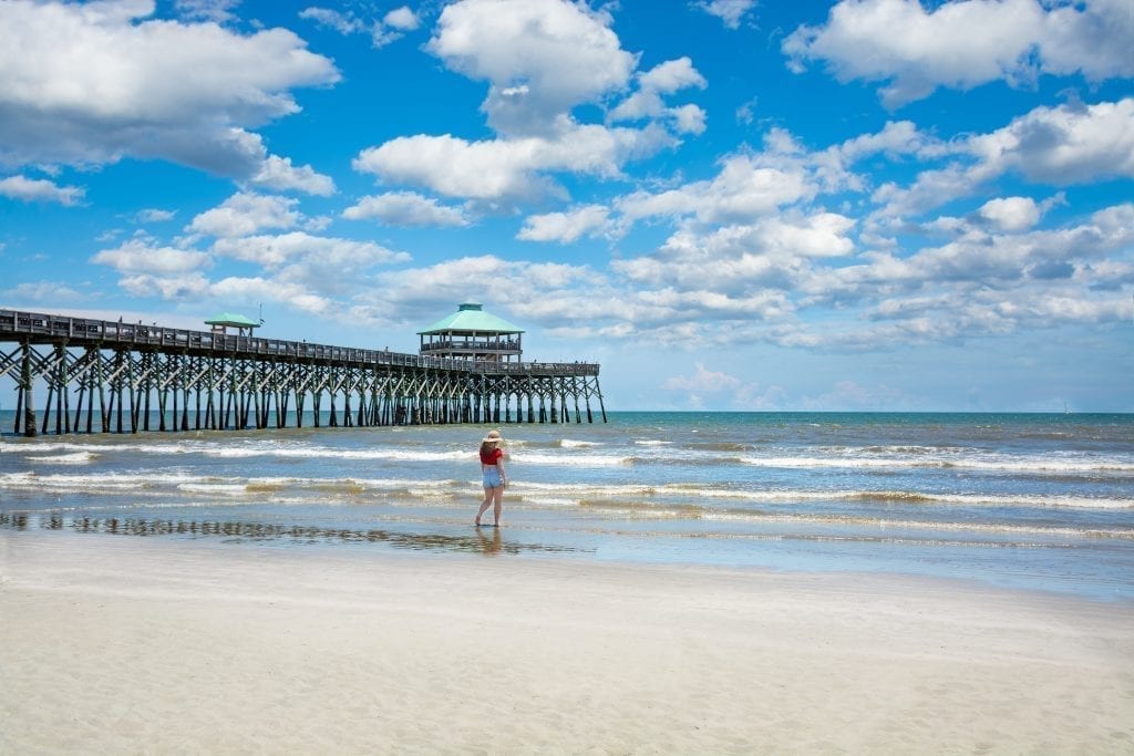 Woman walking along the water next to the pier at Folly Beach on a partly cloudy day. Folly Beach is a fun place to visit during a weekend in charleston sc