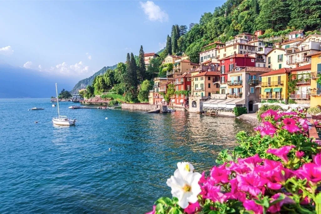 lake como with a sailboat on it with the village of varenna visible to the right side of the photo