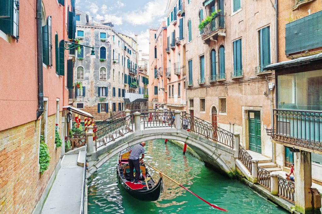 venetian canal with a gondola passing under a bridge. one of the fun facts about venice italy is that there are far fewer gondolas now than in the past
