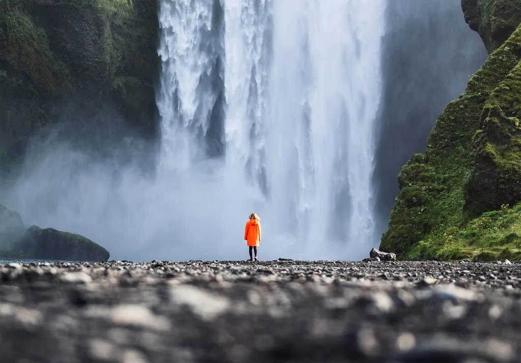 woman in an orange raincoat standing in front of skogafoss waterfall in iceland, one of the best tourist attractions in europe