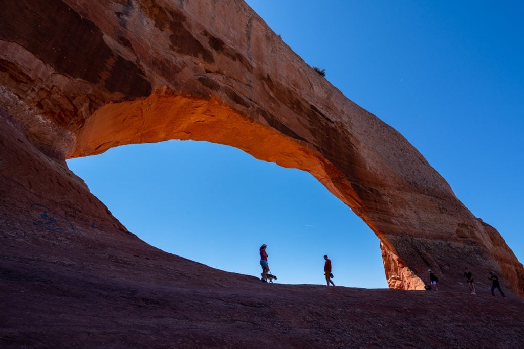 view of hikers standing inside wilson arch moab utah with blue sky behind them