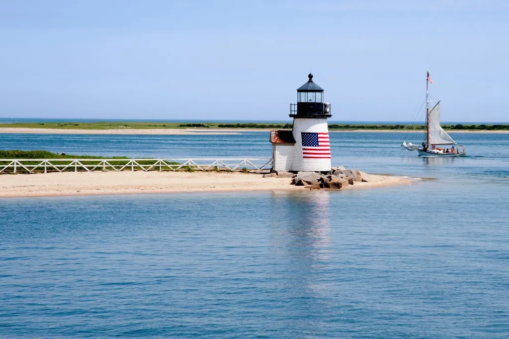 white lighthouse with an american flag on it and a sailboat nearby in nantucket massachusetts, one of the best tourist attractions in usa