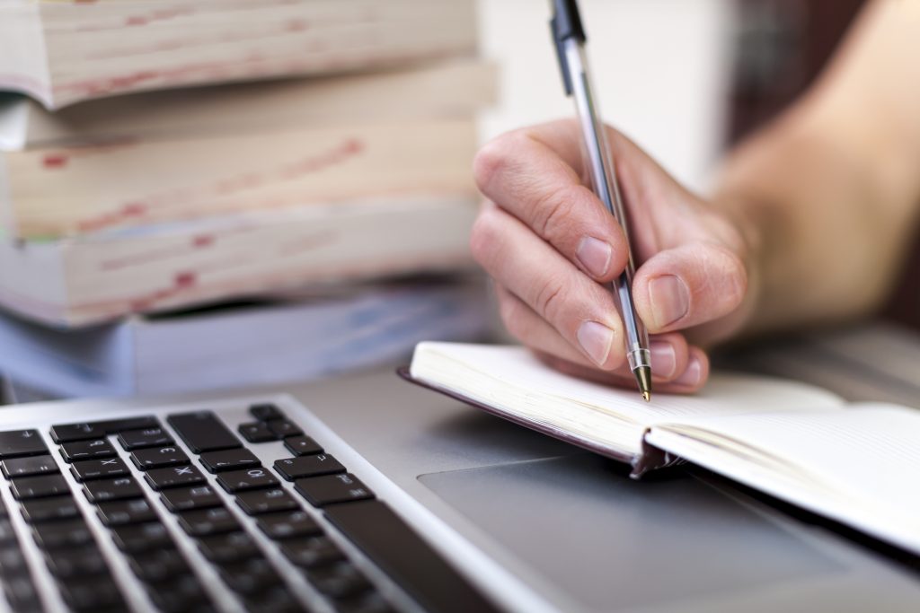 person writing travel journal examples in a notebook with laptop open