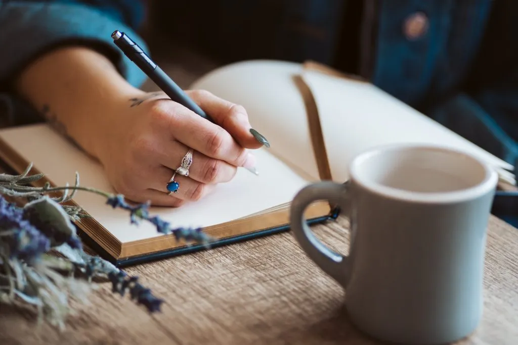 woman writing a travel diary using travel journal prompts at a table with coffee and flowers