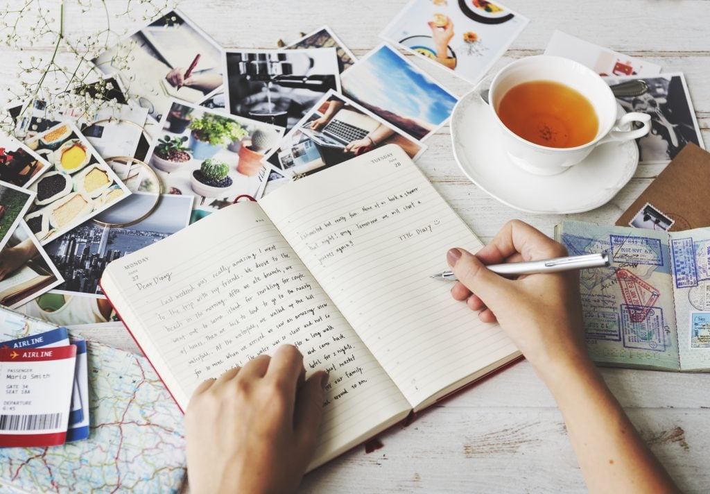 person writing in one of the best travel journals with photos and a cup of tea spread out next to them