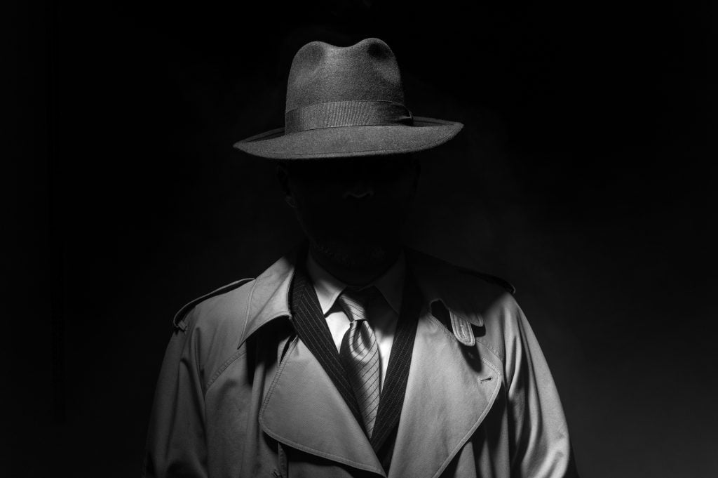 old fashioned mobster in a fedora and trenchcoat with his face hidden in a black and white photo