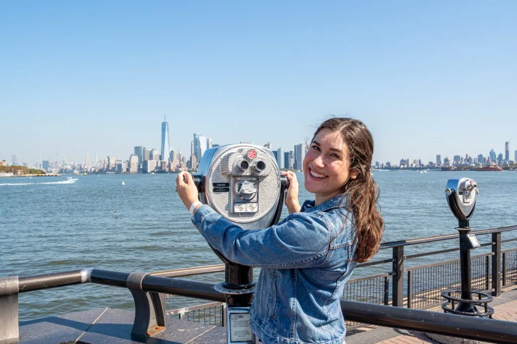 kate storm standing at a magnifying glass on liberty island overlooking financial district manhattan