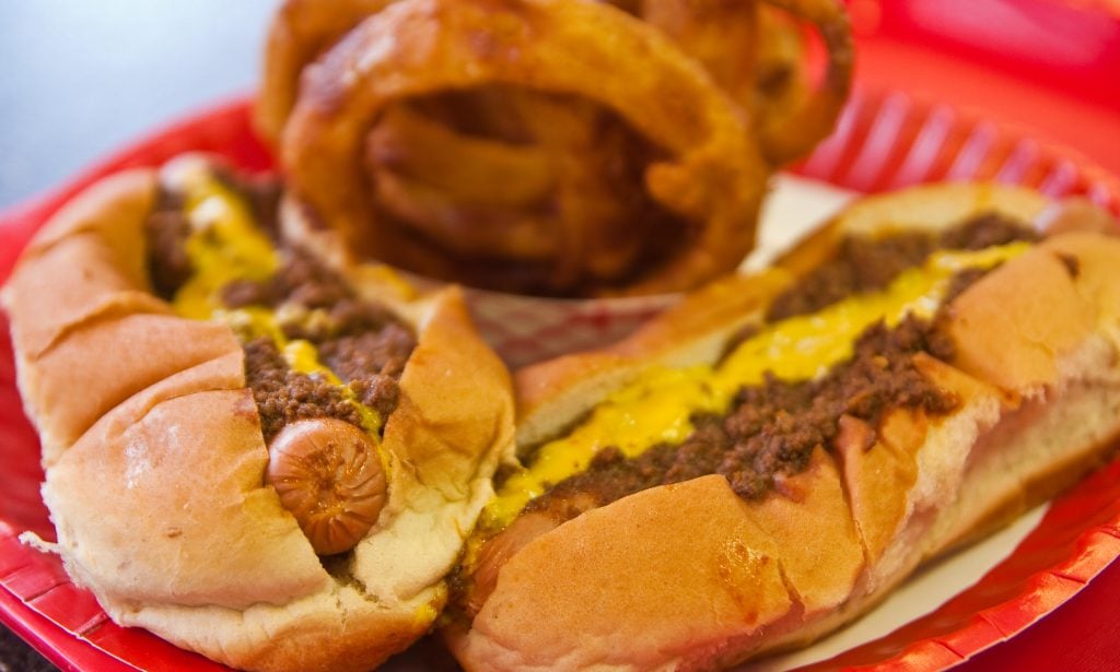 two chili dogs with mustard and onion rings in the background