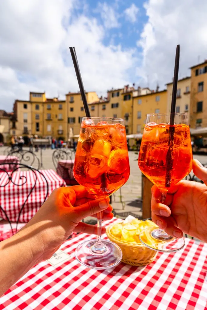 two aperol spritzes being held up in piazza dell'anfiteatro, one of the best places to visit lucca italy