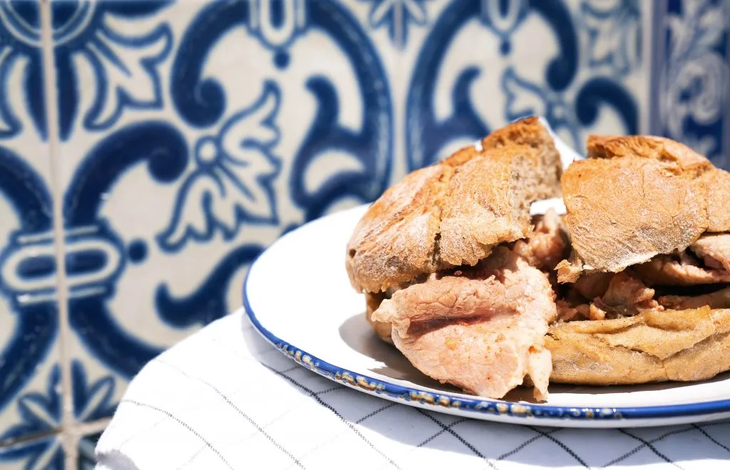 bifana sandwich on a white plate in front of azulejos in portugal