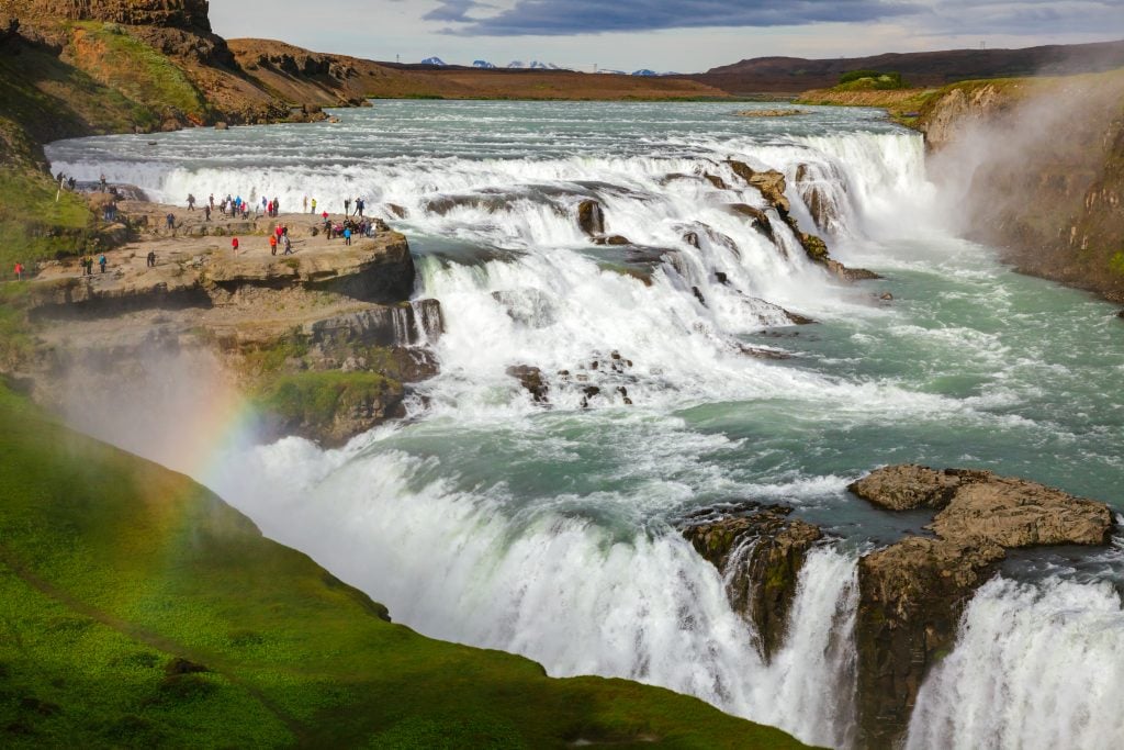 visitors admiring gullfoss with a rainbow in the foreground