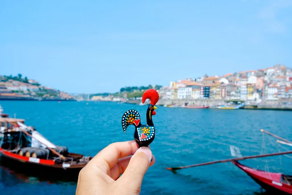 barcelos rooster being held up in front of douro river in porto, one of the best portugal souvenirs