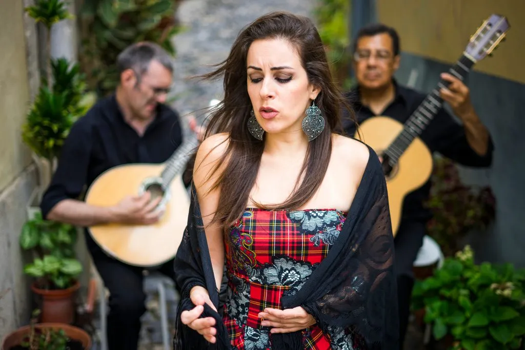 woman singing portuguese fado music in a plaid dress with two men playing guitars behind her